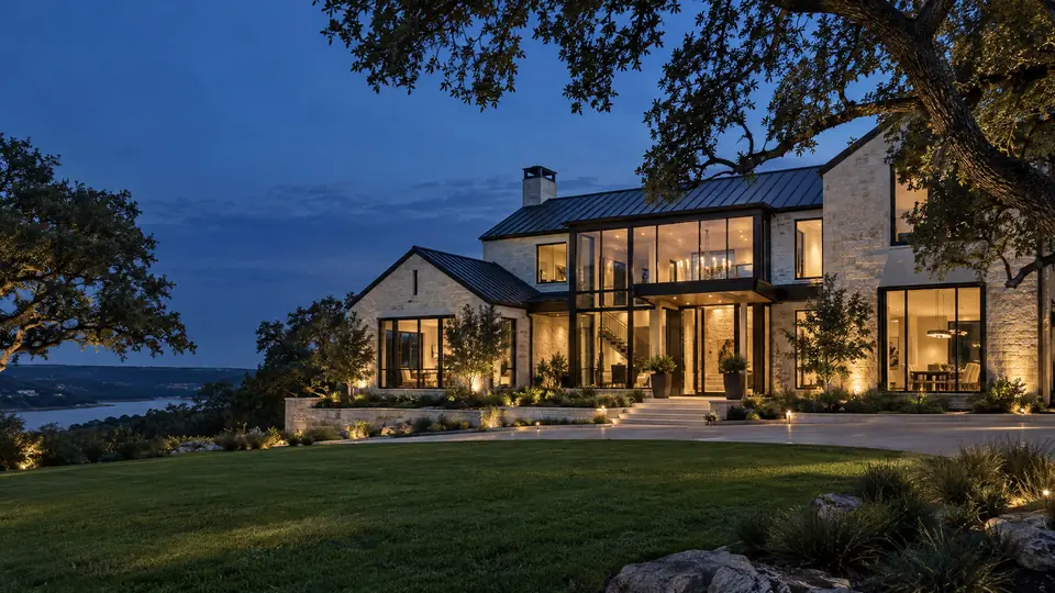 Modern luxury Texas custom home with limestone walls, metal roof and glass walls at dusk under live oaks near Houston, TX